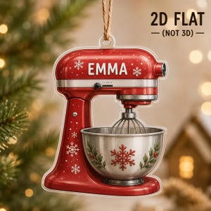 May include: A flat, 2D red stand mixer ornament featuring the name EMMA in white text, decorated with white snowflakes and a silver mixing bowl with a red snowflake and holly leaf design. The ornament hangs from a brown string.