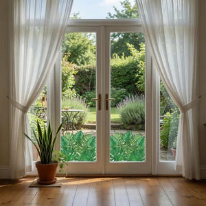 May include: A set of glass door window decals featuring a repeating pattern of green monstera leaves. The decals are applied to the lower panels of a double glass door, which is framed by white curtains and overlooks a green garden.