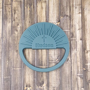 May include: A teal-colored, round, personalized item with a handle. The design features a cross above the name "Hudson" and radiating lines, resembling a sunrise. The item is made of a soft material.