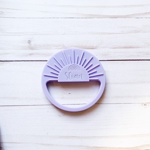 May include: A purple silicone teether shaped like a sun with a rainbow and the name "Sloan" inscribed on it.