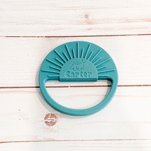 May include: Teal silicone teether with a sunburst design and a dinosaur graphic. The teether has a handle and the name "Carter" is printed on the front.