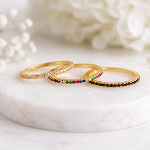 May include: Three thin gold-tone metal rings, each featuring a row of small, round-cut stones. One ring has clear stones, one has a multi-coloured array of red, green, and blue stones, and the third ring features a row of solid black stones.