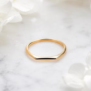 May include: A thin gold ring featuring a smooth, polished finish and a subtle geometric point at the top. The band is circular with a flat, angled section that creates a distinct chevron-like shape, reflecting light across its metallic surface.