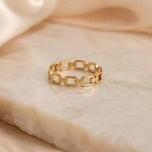 May include: A gold-toned ring featuring a continuous chain-link design. The band is composed of small, uniform, rectangular metal links connected in a repeating pattern, creating a structured and polished appearance for this finger accessory.