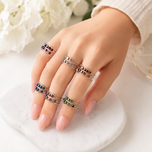May include: A collection of five silver-toned wire rings, each featuring a row of small, square-shaped beads in various colours including purple, blue, green, and metallic brown, designed to wrap around the finger with a flexible, open-ended band.
