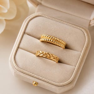 Puede incluir: Dos anillos dorados con una textura de cuerda trenzada o retorcida. El anillo superior presenta un diseño de cuerda más grueso y pronunciado, mientras que el anillo inferior tiene un patrón tejido más fino y delicado. Ambos anillos están en una caja.