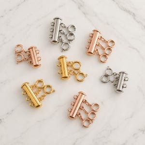 May include: A collection of multi-strand jewellery clasps in silver, gold, and rose gold tones. Each clasp features a cylindrical bar with multiple attached jump rings, designed to connect several strands of necklaces or bracelets together securely.