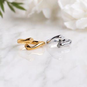 May include: Two open-ended rings, one gold-toned and one silver-toned, each featuring a design of two hands reaching towards each other to form a wrap-around hug shape. The bands are smooth and polished, with the hand details clearly visible.
