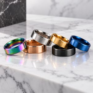 May include: A collection of six metal rings featuring a flat band design with beveled edges. The rings are finished in various colours including iridescent rainbow, rose gold, brushed silver, polished gold, matte black, and deep blue.