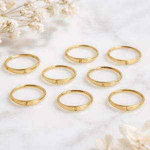 Puede incluir: Una colección de ocho anillos finos de metal en tono dorado, cada uno con una letra grabada. Las letras visibles en los anillos son A, M, S, R, H, F, D y L. Los anillos tienen un acabado liso y pulido con un diseño de banda circular simple.