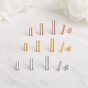 May include: A collection of stud earrings arranged in three horizontal rows by colour, featuring rose gold, yellow gold, and silver tones. Each earring consists of two parallel vertical bars joined together, with post-back closures visible.