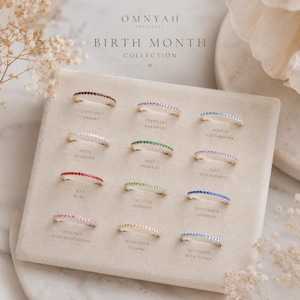 May include: A selection of gold-toned birthstone rings from the "Birth Month Collection" by Omnyah Jewellery. Each ring is set with a row of gemstones, including garnet, amethyst, aquamarine, diamond, emerald, ruby, peridot, alexandrite, sapphire, pink tourmaline, citrine, and blue topaz.