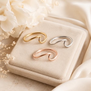 May include: Three rings with a wave-shaped design, each featuring a smooth, continuous band. One ring is gold-toned, one is silver-toned, and one is rose gold-toned. The rings are arranged in a triangular formation on a square, beige velvet display.