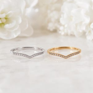 Pode incluir: Dois anéis em forma de chevron, um em metal prateado e outro em metal dourado, cada um com uma fila de pequenas pedras claras de corte redondo ao longo da banda em V. Os anéis estão posicionados lado a lado sobre uma superfície clara.