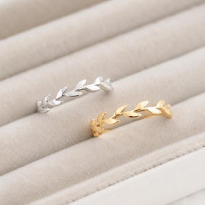 Könnte beinhalten: Zwei dünne Metallringe, einer in Silberoptik und einer in Goldoptik, mit einem durchgehenden Banddesign aus kleinen, polierten Blättern in Rankenform. Die Ringe liegen nebeneinander und zeigen ihre glänzende metallische Oberfläche.