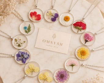 May include: A collection of silver necklaces with round pendants, each containing a pressed flower in various colors. The necklaces are arranged in a circle around a card with the text "OMNYAH JEWELLERY". The flowers include red, purple, yellow, and white blooms.