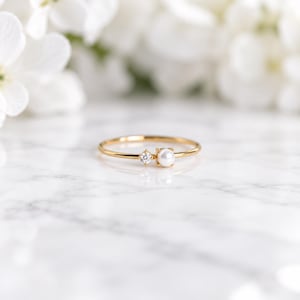Può includere: Un sottile anello in oro con una piccola perla bianca rotonda e una singola pietra preziosa trasparente a taglio rotondo, posizionate fianco a fianco su una delicata fascia in oro lucido. L'anello è posizionato orizzontalmente, evidenziando il contrasto.
