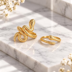 May include: Two gold-toned rings shaped like snakes. The larger ring features a coiled snake with a textured, scale-like surface. The smaller ring is a simple, open-ended band designed to resemble a slender snake with a textured finish.