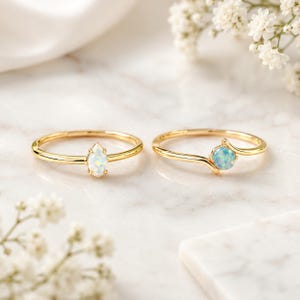 Peut inclure: Deux bagues en or ornées d'opales irisées. La bague de gauche présente une opale taille marquise sur un anneau simple, tandis que la bague de droite possède une opale ronde sur un anneau doré incurvé qui entoure légèrement la pierre.