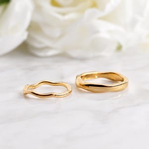 May include: Two gold-toned rings with a smooth, undulating, wavy surface texture. The rings are circular with a polished finish, featuring a continuous, irregular, organic shape that creates a soft, flowing appearance across the metal bands.