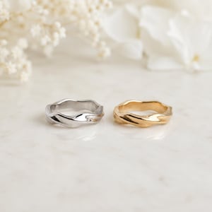Op de afbeelding: Twee ringen met een golvend, onregelmatig bandontwerp. De ene ring heeft een gepolijste zilverkleurige afwerking, terwijl de andere ring een warme, gepolijste goudkleur heeft. Beide ringen hebben een vloeiende, continue en organische vorm.