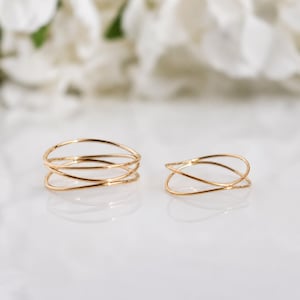 May include: Two gold-toned rings with a thin, delicate band design. The ring on the left features a multi-layered, crisscrossing wire structure, while the ring on the right consists of a single, simple crossover loop design in a polished finish.