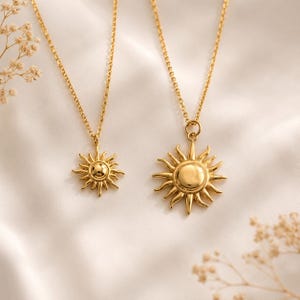 May include: Two gold-toned necklaces featuring sun-shaped pendants with radiating rays. The pendants are suspended from delicate gold-toned chains. The sun designs have a smooth, polished centre and pointed rays extending outward in a circular pattern.
