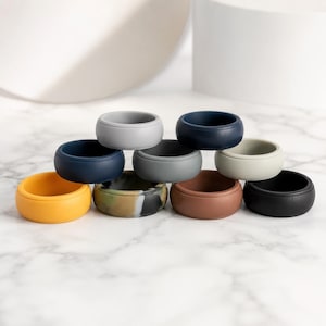 May include: A collection of nine silicone rings in various solid colors including yellow, navy blue, light gray, dark gray, off-white, brown, and black, along with one ring featuring a marbled pattern of green, black, and white tones.