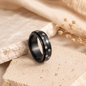 May include: A black ring featuring a central spinning band with a series of evenly spaced, vertical silver-toned metallic rectangular accents. The ring has a smooth, matte finish and a consistent width throughout its circular structure.