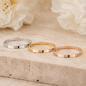 May include: Three thin metal rings in silver, yellow gold, and rose gold tones. Each ring features a smooth, flat band design with three small, round, clear stones set at equal intervals along the outer surface of the circular metal bands.