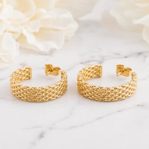 May include: A pair of gold-toned hoop earrings featuring a textured, woven-link chain design. Each earring is constructed as a semi-circular hoop with a post-back closure mechanism, showcasing a consistent metallic finish across the entire surface.