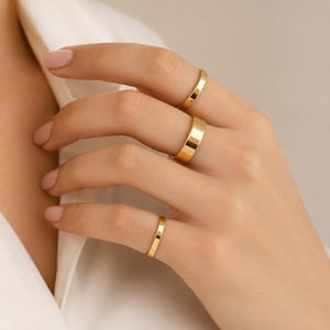 Puede incluir: Tres anillos de oro con un acabado liso y pulido y un perfil de banda plano. Los anillos se llevan en el dedo índice, medio y anular, mostrando un diseño sencillo y uniforme con una superficie reflectante que capta la luz.