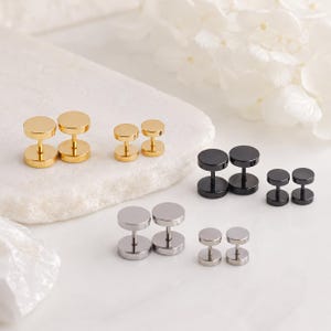 May include: A collection of six pairs of barbell-style stud earrings in three distinct metallic finishes. The set includes two pairs of gold-tone, two pairs of black, and two pairs of silver-tone earrings, each featuring a circular flat front and back.