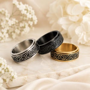 Peut inclure: Trois bagues en métal présentant un motif répétitif de nœuds triquetra celtiques. Une bague est argentée, une est noir mat et une est dorée. Chaque anneau a un profil large et plat avec le motif de nœud gravé sur toute la surface extérieure.