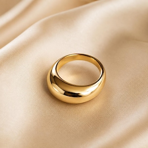 May include: A polished gold-tone ring with a thick, rounded, and smooth band. The ring features a continuous, solid metal construction with a high-shine reflective surface that catches light, creating a bold and substantial appearance on the finger.