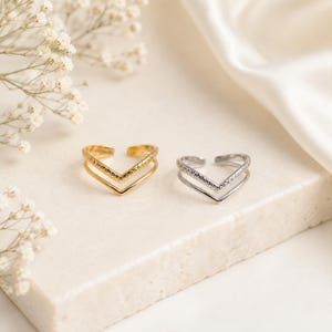 May include: Two open-ended chevron rings, one in a polished gold tone and one in a polished silver tone. Each ring features a double-band design where the top band has a textured, cross-hatched pattern and the bottom band is a smooth, plain wire.