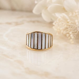 May include: A gold-toned ring featuring a series of vertical, rectangular white mother-of-pearl inlays arranged side-by-side across the front of the band. The metal frame holds each individual inlay securely, creating a structured, multi-panel design.