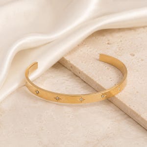 May include: A gold-toned cuff bracelet featuring a polished metal band with evenly spaced star-shaped engravings. Each star motif is centred with a small, clear, sparkling stone, creating a consistent pattern along the outer surface of the open-ended bangle.