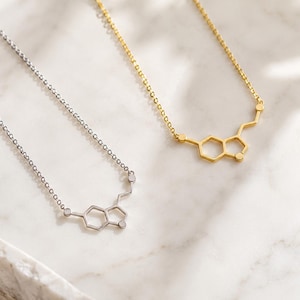 May include: Two necklaces featuring a serotonin molecular structure pendant. One necklace is finished in a polished silver tone, while the other is finished in a gold tone. Both pendants are attached to delicate, thin link chains.