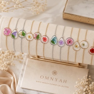 Peut inclure: Une collection de bracelets délicats avec des fleurs pressées encapsulées dans des pendentifs transparents ronds ou en forme de goutte. Les bijoux sont présentés sur un support beige avec le texte OMNYAH BIRTH MONTH FLOWER COLLECTION en doré.