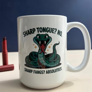 May include: A white ceramic mug featuring a green snake illustration with a red forked tongue. The text reads: SHARP TONGUE? NO. SHARP FANGS? ABSOLUTELY. The mug has a curved handle and is positioned next to a small, dark red lipstick tube.