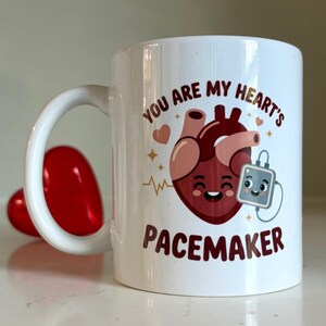 May include: A white ceramic mug featuring a cartoon illustration of a smiling anatomical heart connected to a small pacemaker device. The text reads "YOU ARE MY HEART'S PACEMAKER" in dark brown letters, accented by small hearts and sparkles.