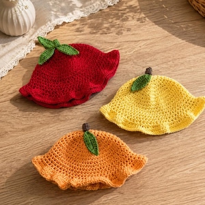 May include: Three crocheted bucket hats in red, yellow, and orange. Each hat features a ruffled brim and a small green leaf accent on top with a brown stem. The red hat has a larger leaf detail, while the yellow and orange hats have single leaves.