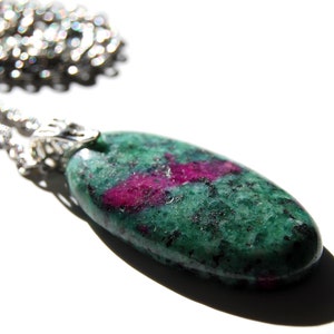 May include: An oval-shaped pendant featuring a polished green zoisite stone with vibrant magenta ruby inclusions, suspended from a silver-toned metal chain. The stone has a smooth, glossy texture with dark mineral flecks throughout the surface.