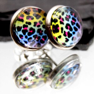 May include: A pair of round stud earrings featuring a vibrant rainbow leopard print pattern with yellow, blue, and pink spots set under a clear glass dome. The earrings are encased in a polished silver-toned metal frame with a reflective finish.