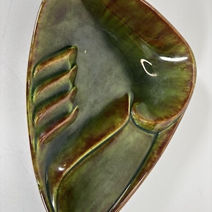 May include: A triangular ceramic ashtray featuring a glossy glaze in shades of olive green and deep brown. The piece has a smooth, curved interior with three molded cigarette rests along one side and a deep, rounded basin for ash collection.