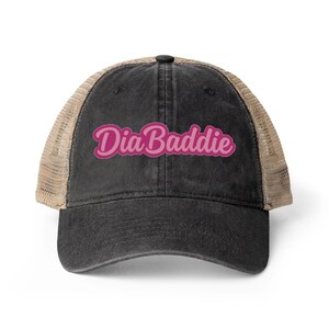 May include: A black trucker hat with a mesh back and a curved brim. The hat features the pink text "Dia Baddie" across the front. The hat is a casual accessory.