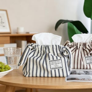 Puede incluir: Dos fundas de tela para cajas de pañuelos con base blanca y rayas verticales azul marino o marrón. Cada funda tiene una parte superior fruncida con un pañuelo blanco y una etiqueta rectangular blanca con el texto MEZO ELEMENT al frente.