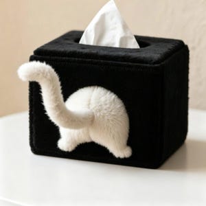 Peut inclure: Une boîte à mouchoirs carrée noire dotée d'une queue et d'un arrière-train de chat en peluche blanche fixés sur la face avant. La boîte est fabriquée dans une matière douce et duveteuse avec une ouverture rectangulaire sur le dessus.