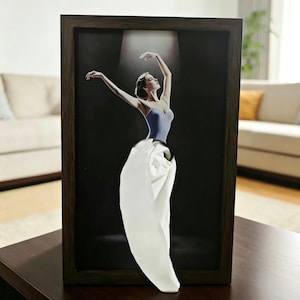 May include: A framed wall art piece featuring a ballerina in a blue leotard and a three-dimensional white fabric skirt that extends outward from the dark background. The artwork is enclosed in a simple, dark brown wooden frame.